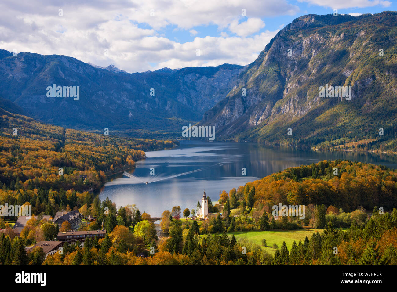A ferry boat towards the town of Ribčev Laz next to Lake Bohinj and trees in autumn colors and Julian Alps mountains in a fall landscape in Slovenia. Stock Photo