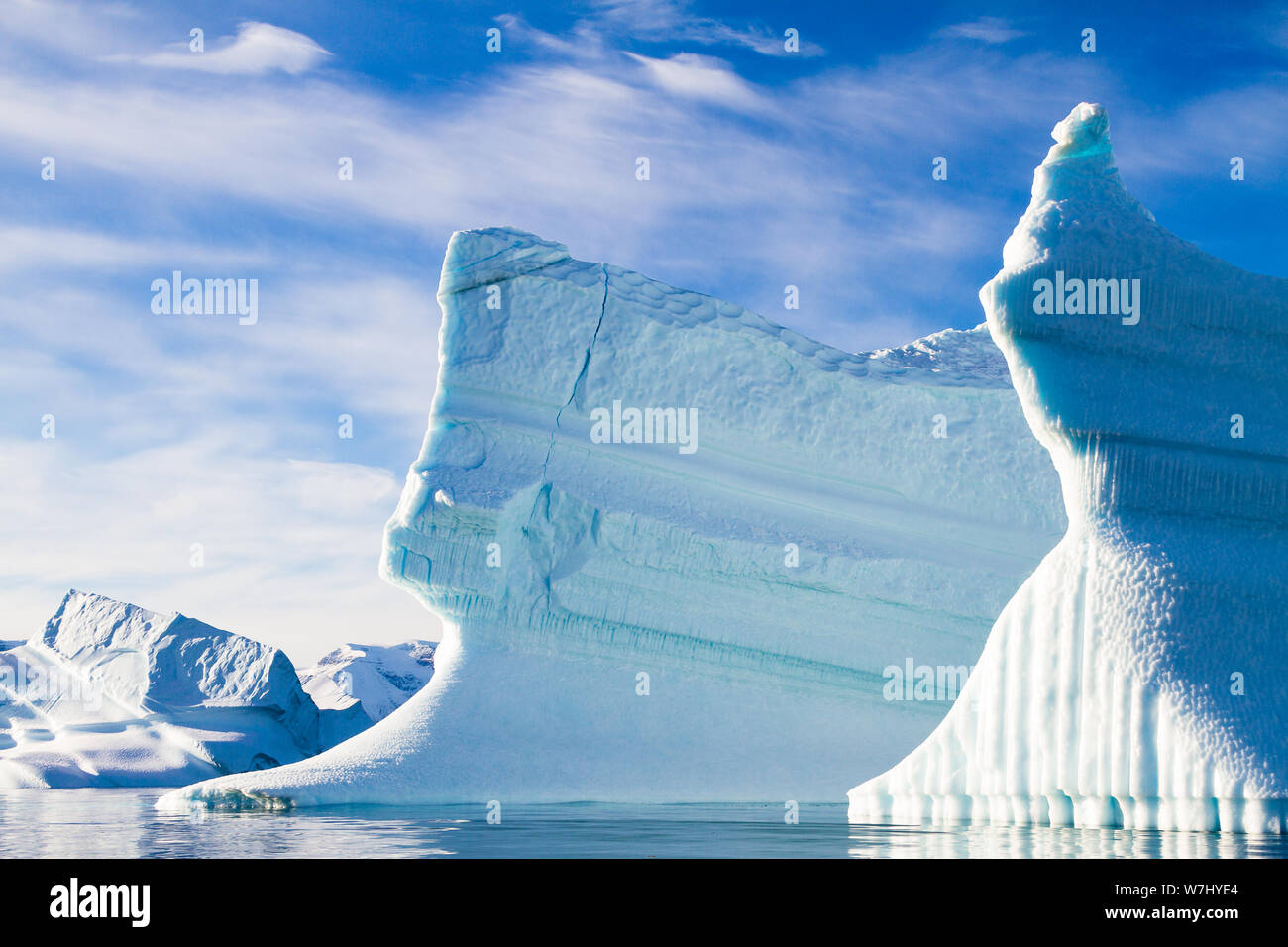 Two towering icebergs stand firmly in the freezing waters of Scoresby Sund, in Eastern Greenland.  Both layered in contrasting ribbons of smooth and fluted ice, the top of the right berg is lit by the sun like a glowing blue torch. Stock Photo