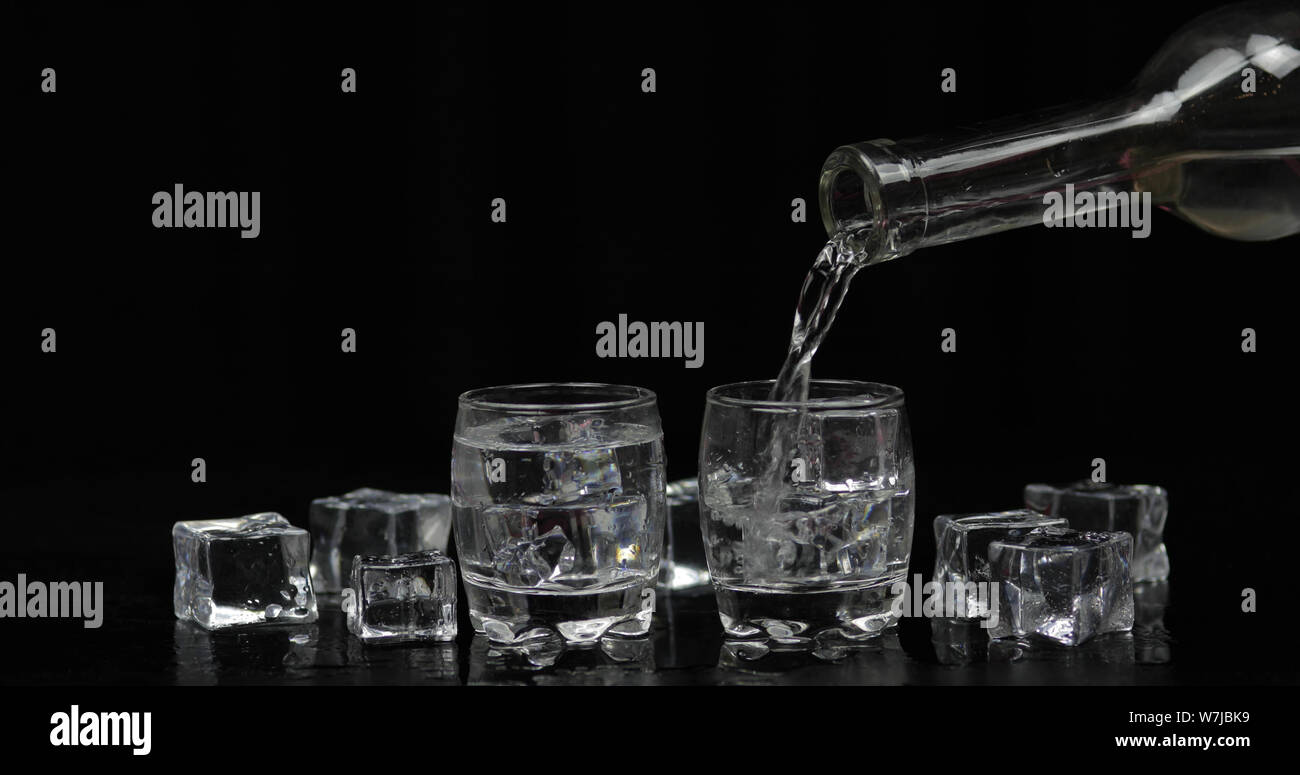 Pouring up shots of vodka from a bottle into drinking glass against black background with ice cubes. Pour of alcohol drink vodka tequila Stock Photo