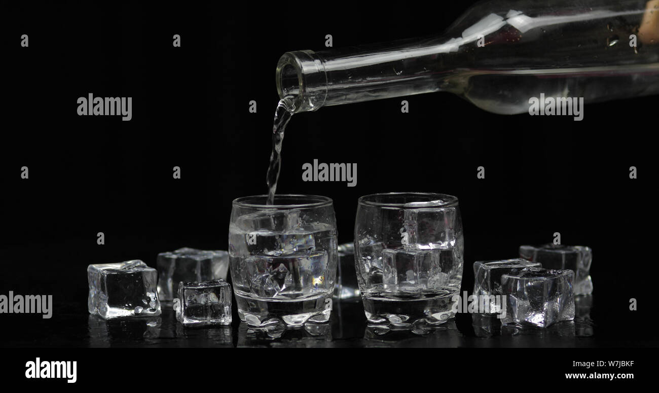 Pouring up shots of vodka from a bottle into drinking glass against black background with ice cubes. Pour of alcohol drink vodka tequila Stock Photo