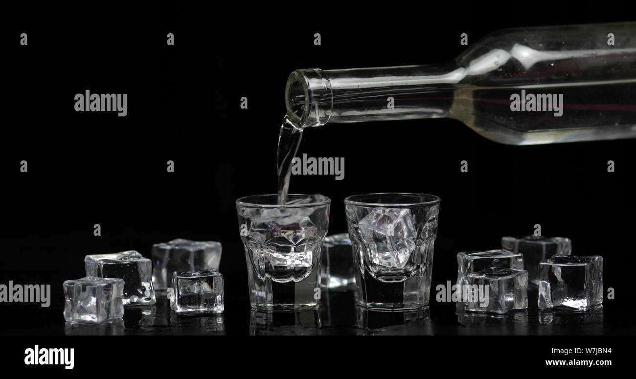 Pouring up shots of vodka from a bottle into drinking glass against black background with ice