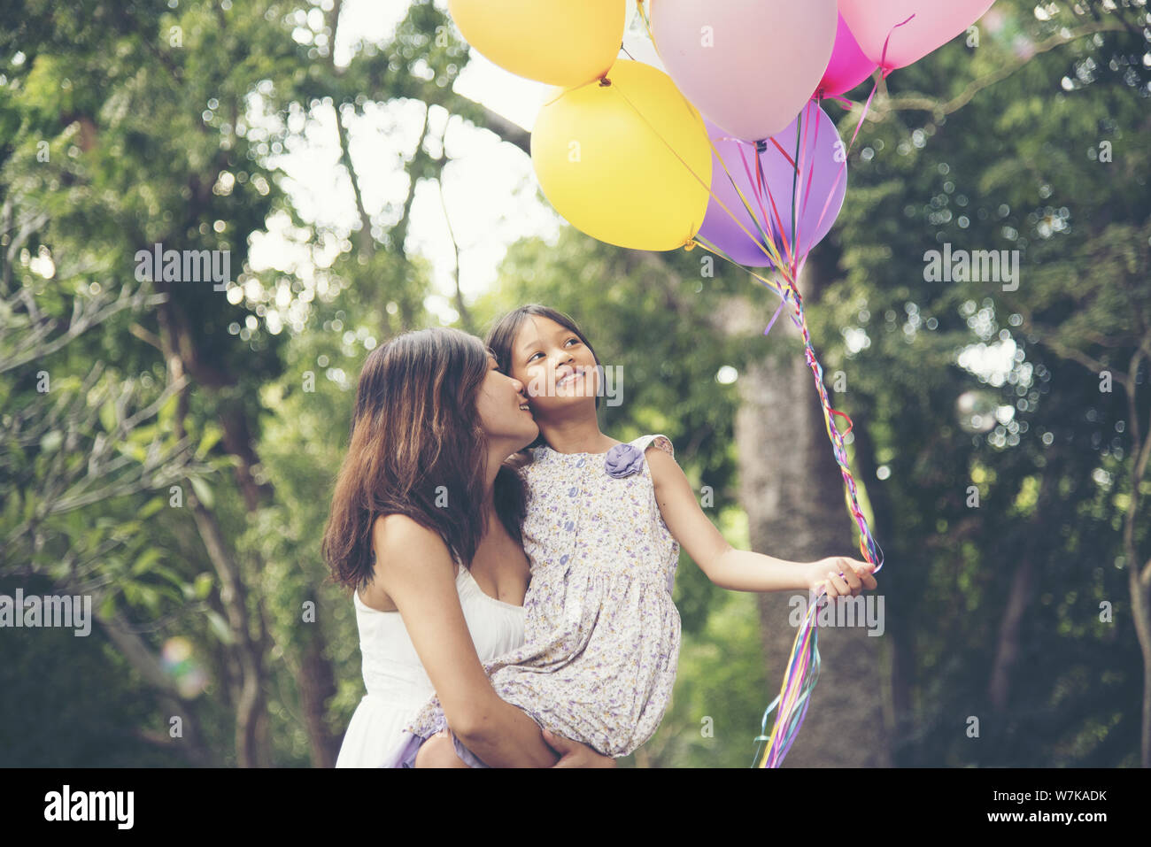 Happy girl holding colorful of air balloons on a green meadow with cloudy and blue sky. Mother hold daughter. Stock Photo
