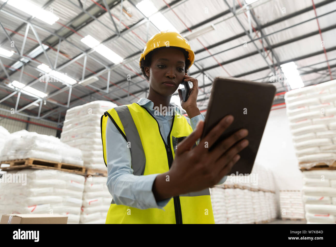 Female worker using digital tablet while talking on mobile phone in warehouse Stock Photo