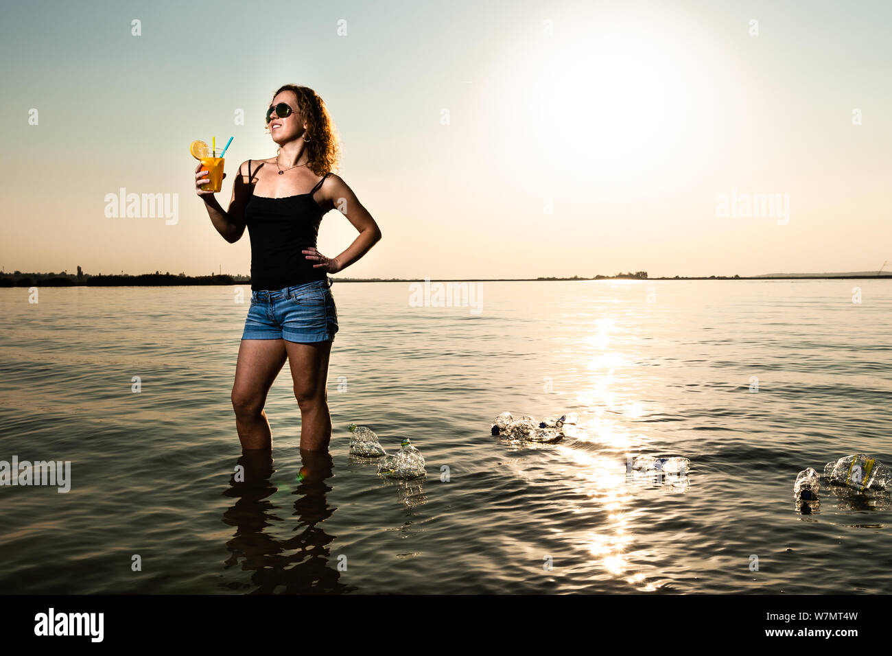 Woman in the sea holding a glass of juice with plastic straw around many plastic bottle swimming on the water. Environmental protection concept. Stock Photo