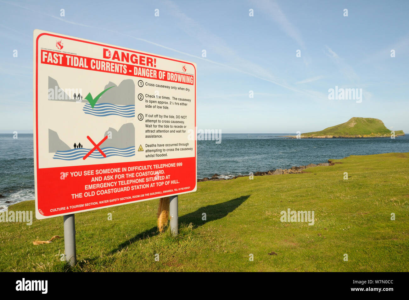 Sign warning when it is unsafe to cross a tidal causeway, with the Worm's head cut off from the mainland as an island at high tide, Rhossili, The Gower peninsula, Wales, UK, July. Sequence 2 of 2 matching view with different tides Stock Photo