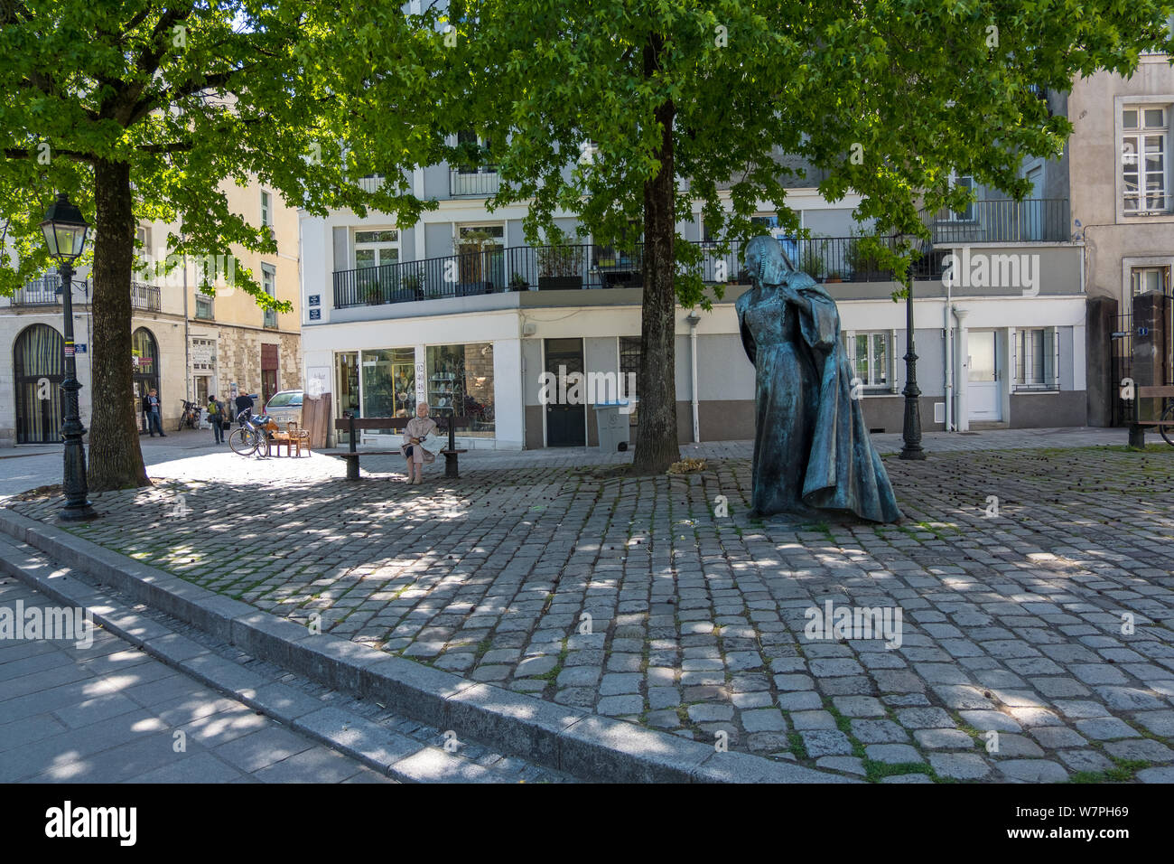 Nantes, France - May 12, 2019: One elderly woman sitting on the bench under green trees near statue of Anne de Bretagne Queen of France. Nantes France Stock Photo