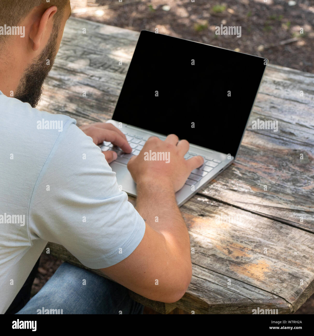 Men's hands with laptop close-up, shoulder shot. Man or guy working on a computer with a blank screen for copy space, sitting on a bench outdoors in a Stock Photo