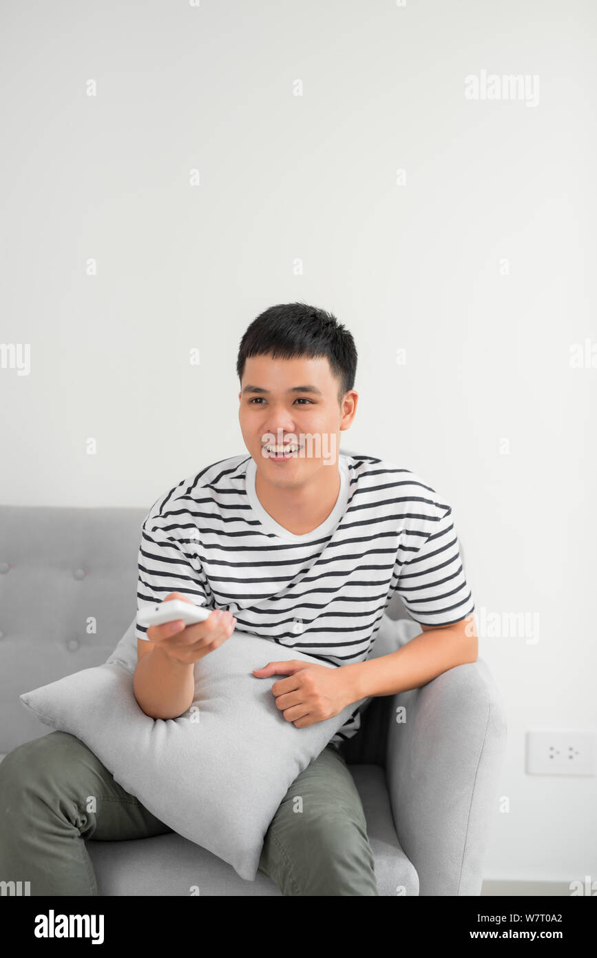 Photo of joyful man 30s in casual clothing sitting on sofa in living room and looking on camera with remote control in hand Stock Photo