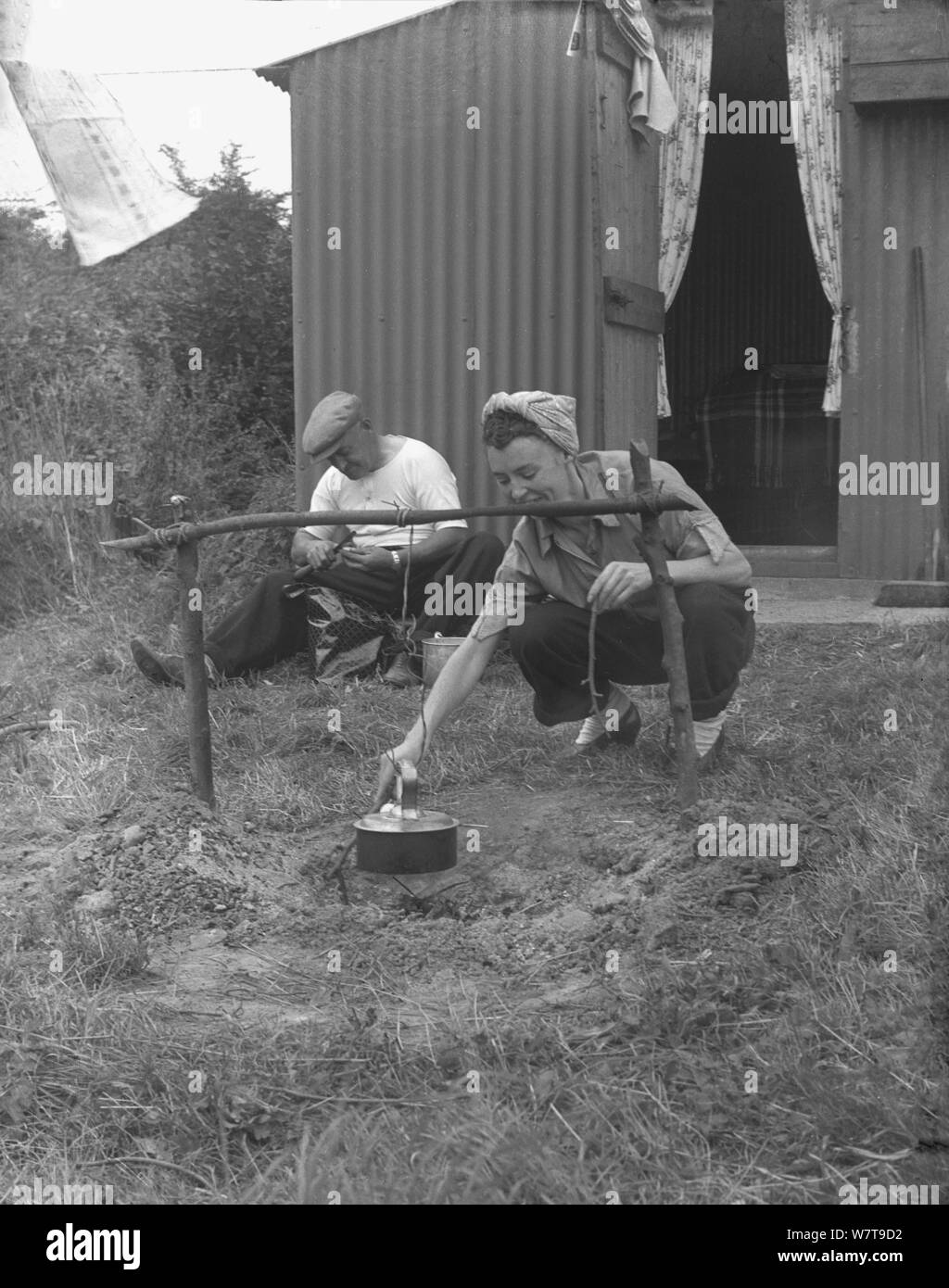 1951, historical, hop picking, an elderly man man and a younger woman outside their hopper hut, with the man peeling potatoes and the lady with a scarf over her hair boling a kettle over a camp fire, Kent, England, UK. The huts, made from corrugated metal (steel) were the basic lodgings for the labourers working in the fields. Stock Photo