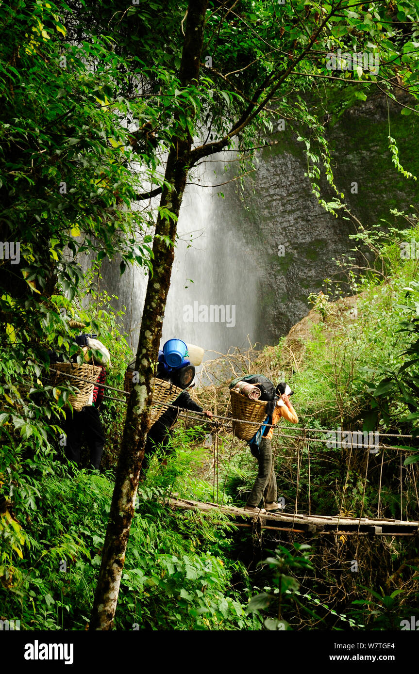 Porters carrying loads across swing bridge  in front of waterfall in the Modi Khola river valley. Annapurna Sanctuary, central Nepal, November 2011. Stock Photo