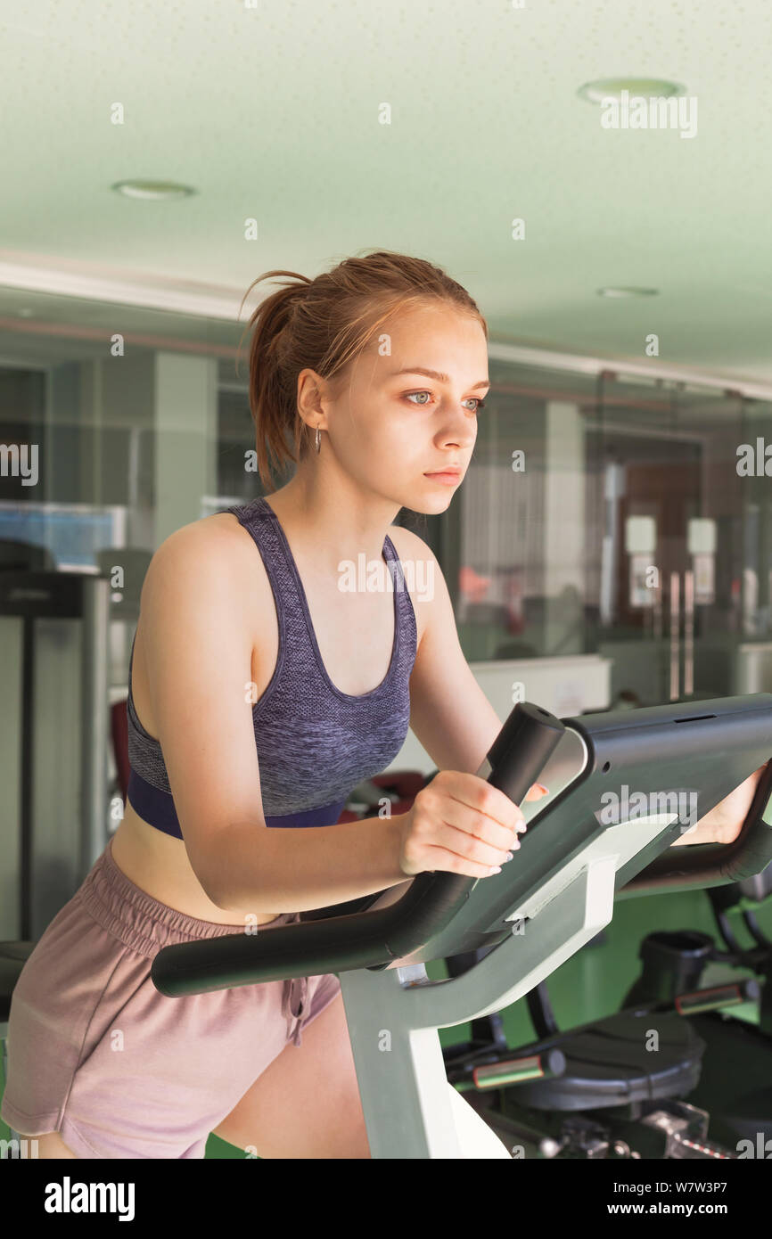 Young beautiful blond sporty girl is engaged on a stationary bicycle in a gym. Vertical photo Stock Photo