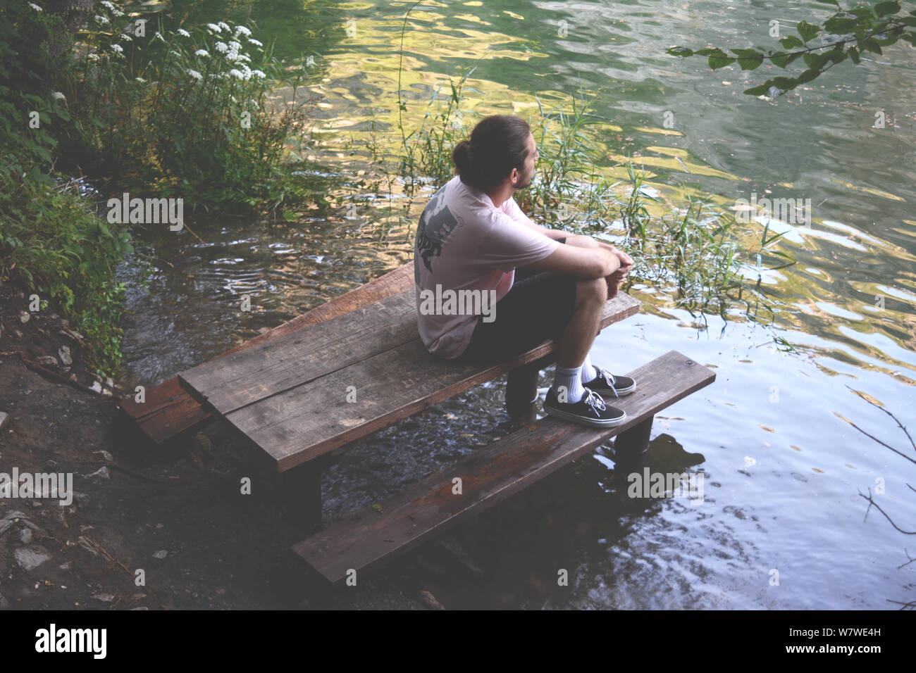 A young man sitting on a flooded wooden bench on a lakeshore. Looking in the distance, contemplating... Stock Photo