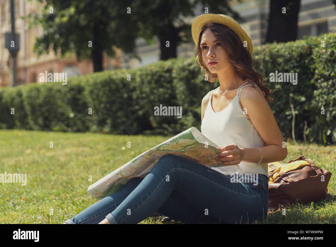 Beautiful young woman is sitting on grass enjoy sunny day in park. Summer happy moment. In search of something on the map. Stock Photo