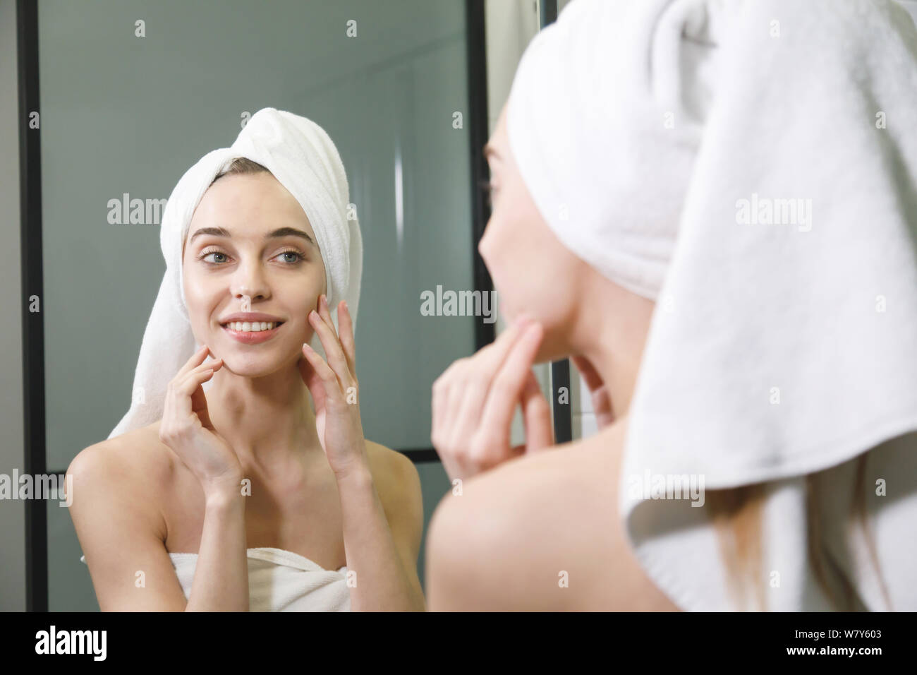 Woman in white bath towel on her head caring of her beautiful skin on the face while standing near the mirror in the bathroom Stock Photo