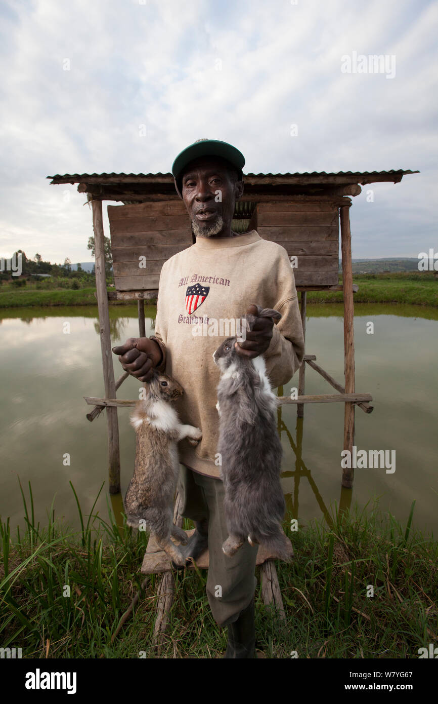 Rwandan farmers with rabbits, outside their hutches which sit over a fish-pond, Rwanda, June 2014. Stock Photo