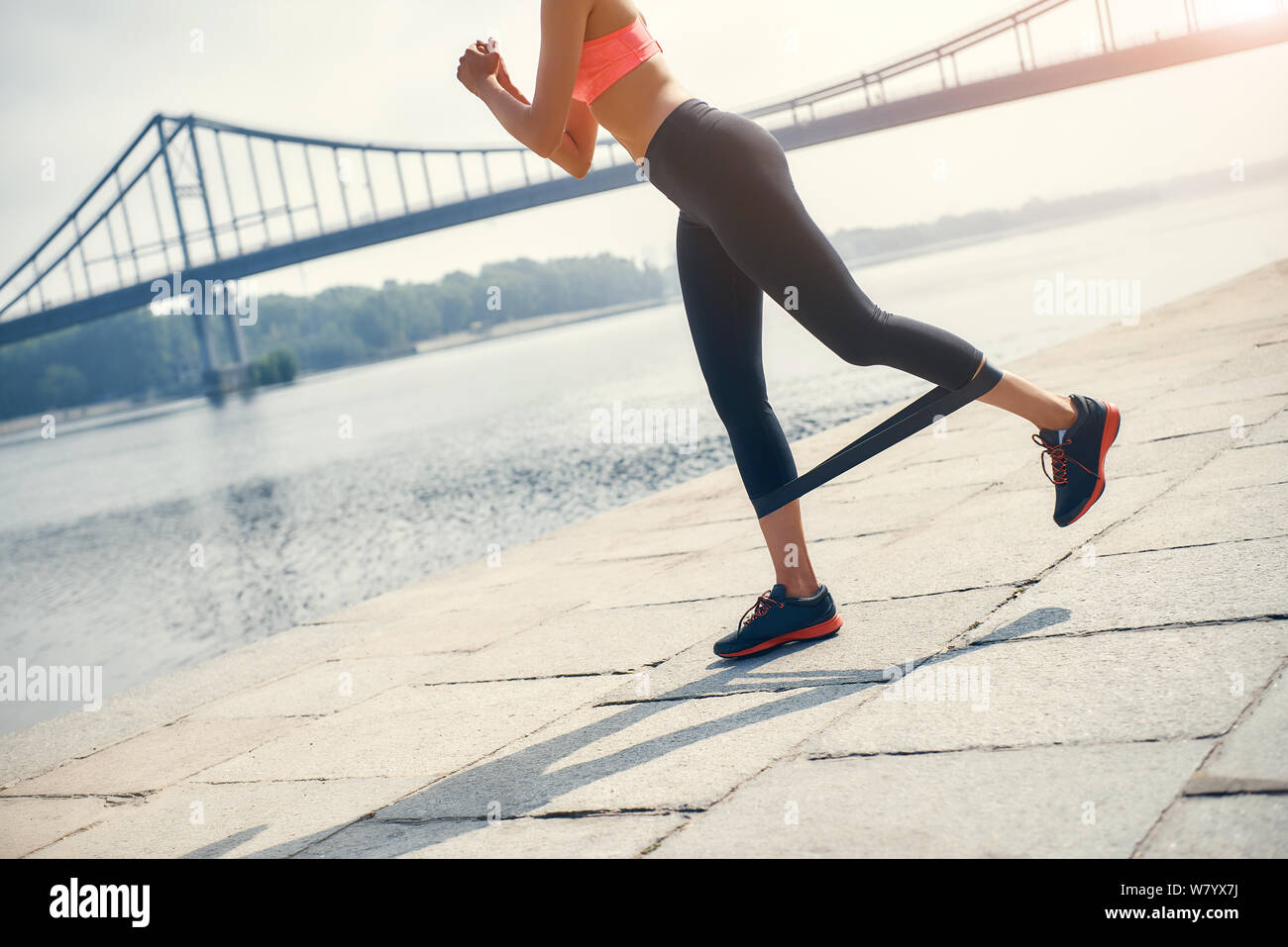 Making perfect legs. Cropped photo of female in sportswear doing exercises with rubber band outdoors in front of the river. Sport concept. Fitness equipment Stock Photo