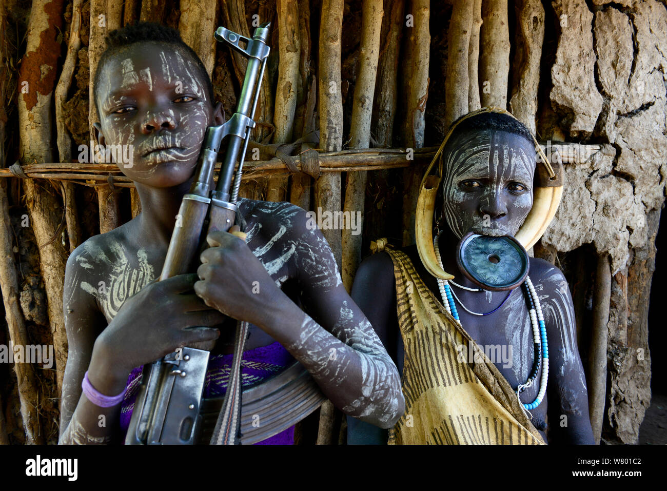 Mursi boy with gun, with mother who has a lip plate, Mursi tribe. Mago National Park. Ethiopia, November 2014 Stock Photo