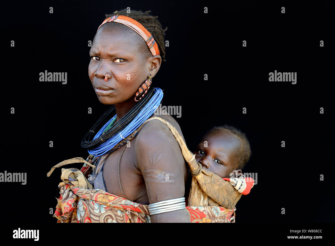 Woman from the Toposa tribe with skin scarifications on face and arms, and wearing traditional head-wear and jewels, carrying her baby on her back, Omo Valley, Ethiopia, March 2015. Stock Photo