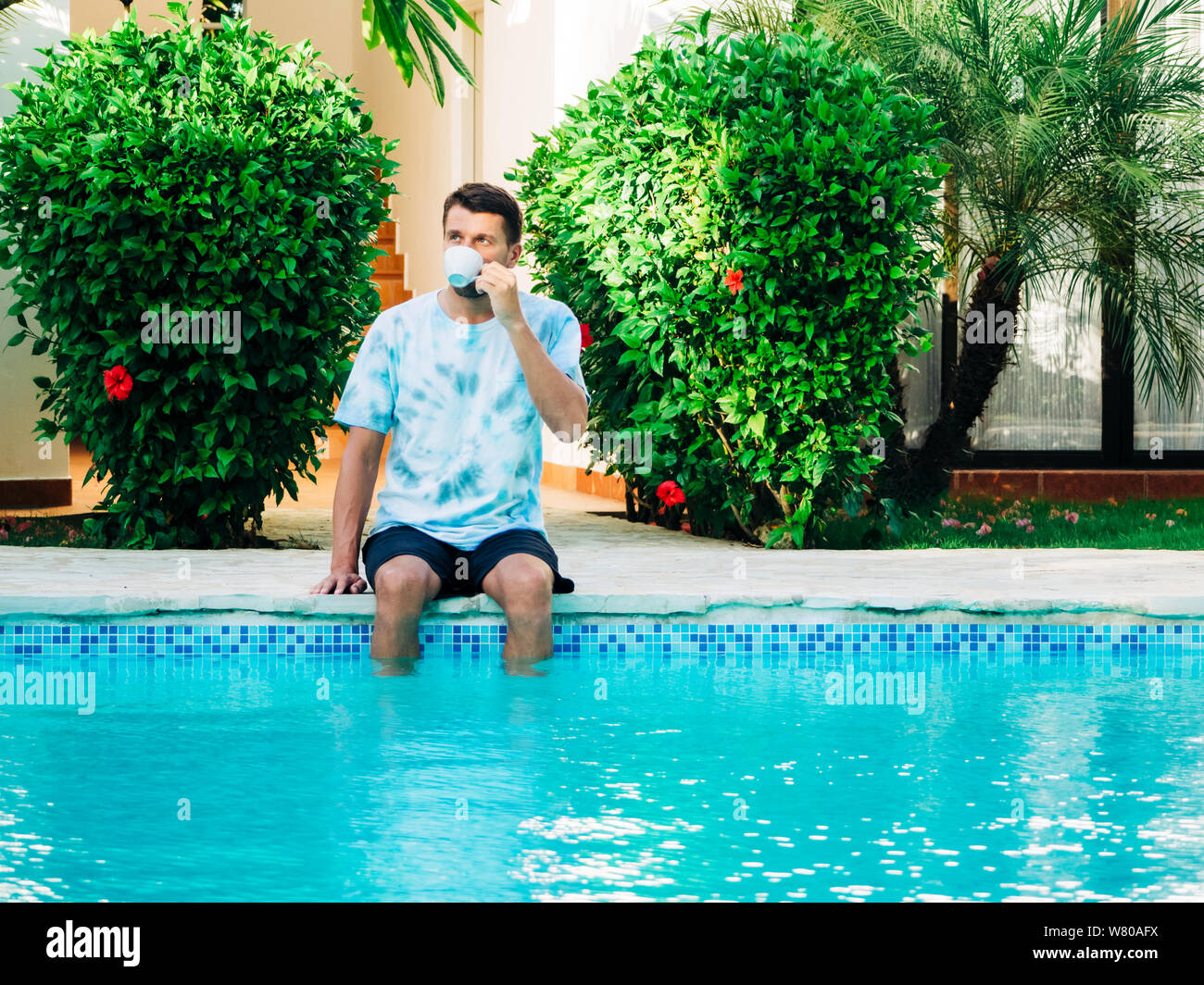 Portrait of a mature age man wearing a t-shirt and shorts sitting near a swimming pool drinking coffee. feet in the pool in the water. Stock Photo