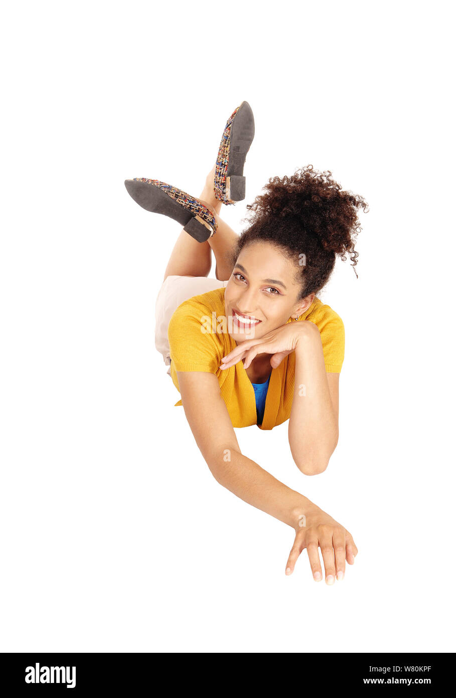 A beautiful multi-racial woman lying on her stomach on the floor in  a yellow sweater and beige shorts with her curly black hair in a bun, isolated fo Stock Photo