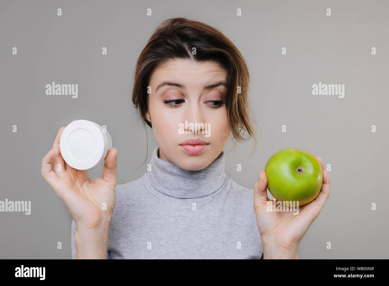 Pretty young woman holding a hyaluronic cream and a apple against a grey background and looking at apple. Lovely girl dressed in grey clothes making c Stock Photo