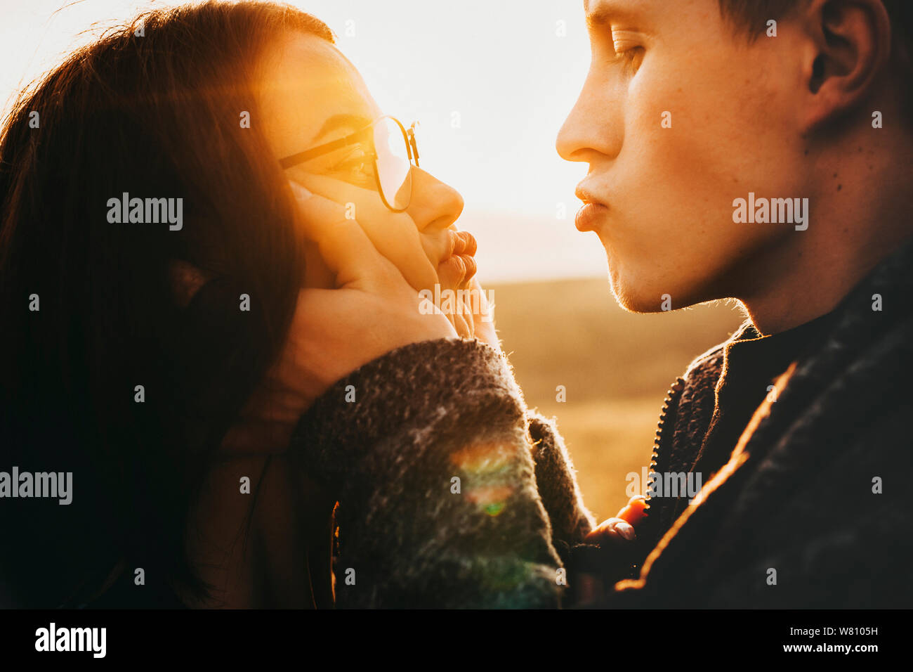 Lovely young couple looking one to another and having fun while man is holding his girlfriend's face in his hands while doing a funny face against sun Stock Photo