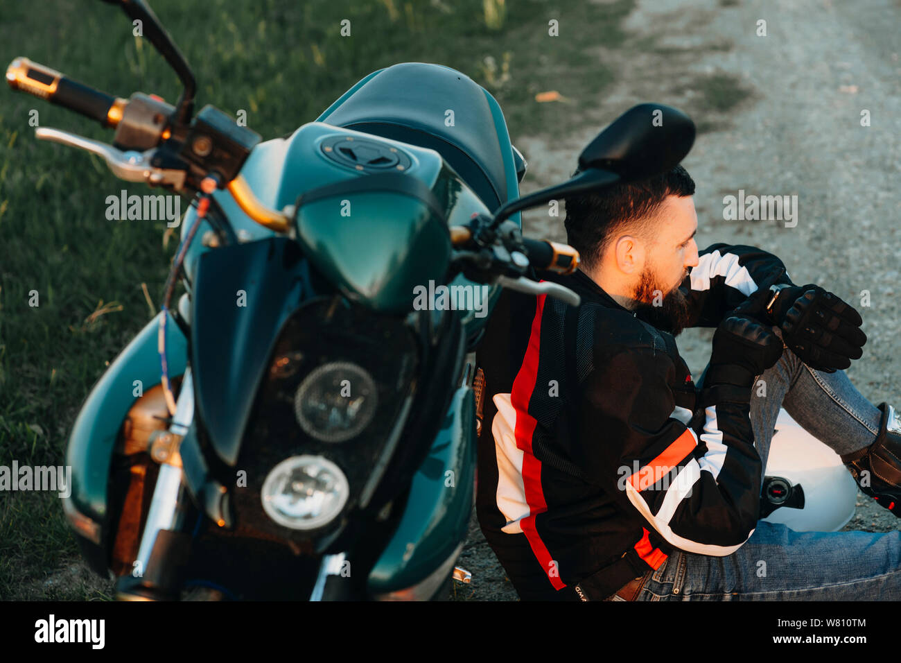 Young bearded biker sitting on the ground and leaning on his bike while ...