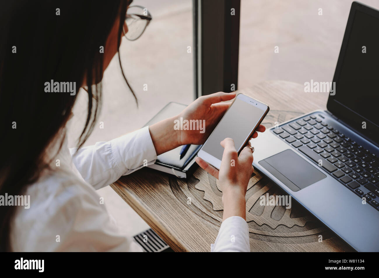 Female hands using a smartphone for operating on internet transaction and laptop sitting at a table in coffee shop. Stock Photo