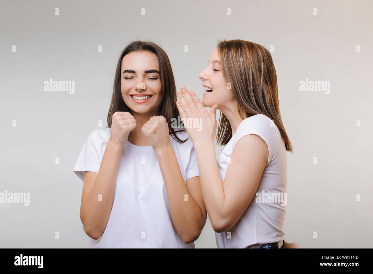 Two lovely women gossiping while one is telling something to another while another is laughing with closed eyes being happy against a grey wall.. Two Stock Photo
