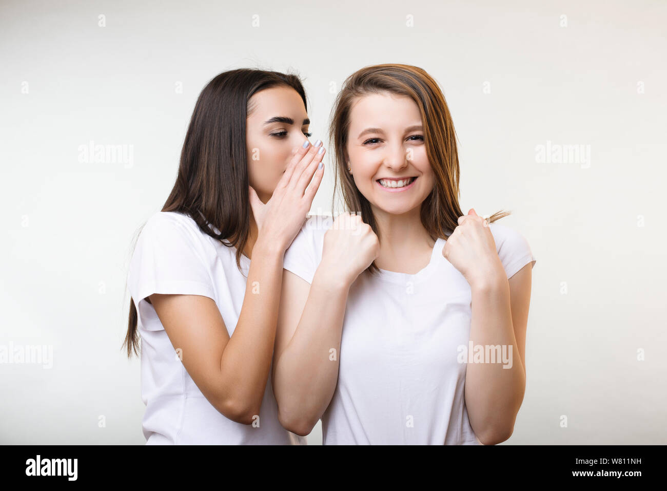 Beautiful young caucasian female telling a secret to her girlfriends ear while she is smiling with closed eyes and tightened punches isolated on a whi Stock Photo