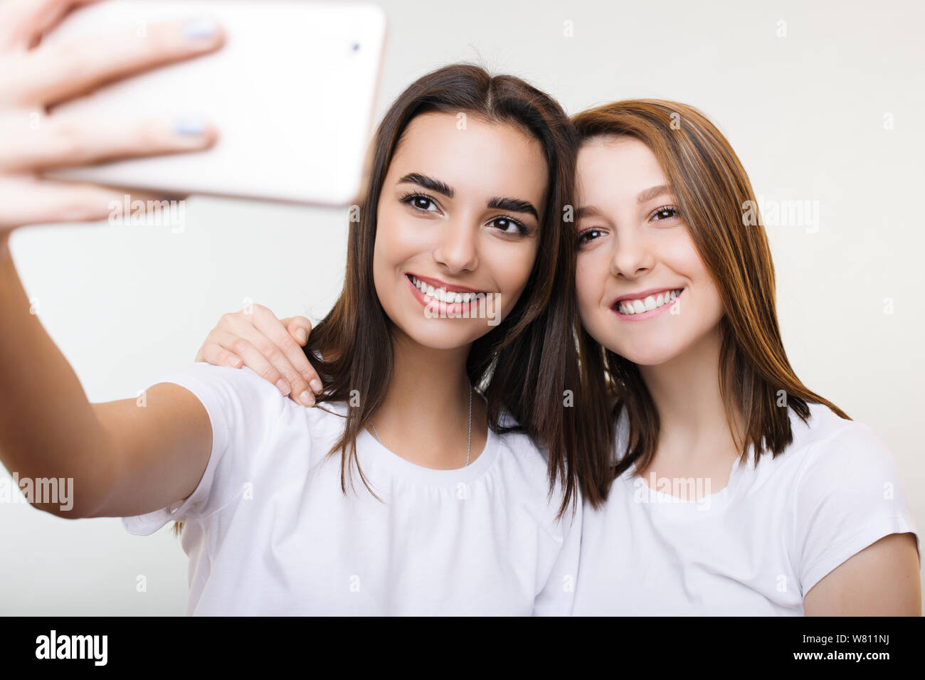 Two beautiful young woman dressed in white doing a selfie smiling and embracing isolated on a white studio background. Stock Photo