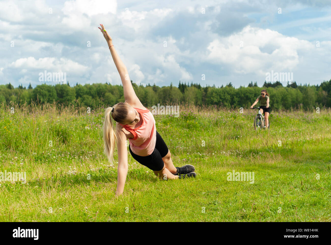 Perm, Russia - July 23, 2019: young woman doing exercise side plank on the grass outdoor while a female cyclist rides in the background Stock Photo