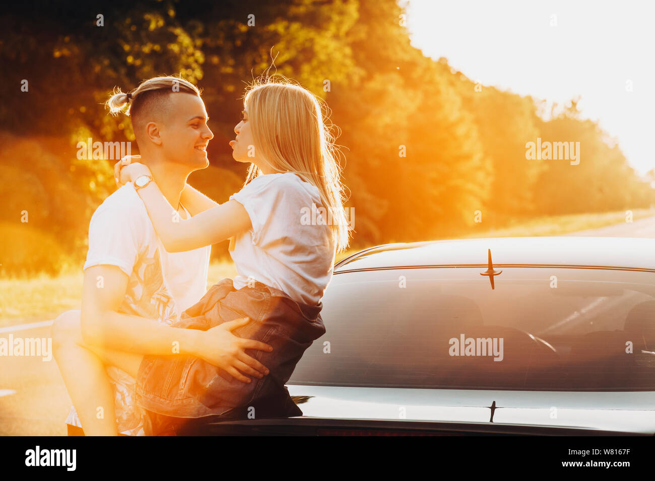 Beautiful young blonde woman having fun with her boyfriend while she is shoing the tongque while he is laughing.Girl sitting on the back of the car wh Stock Photo
