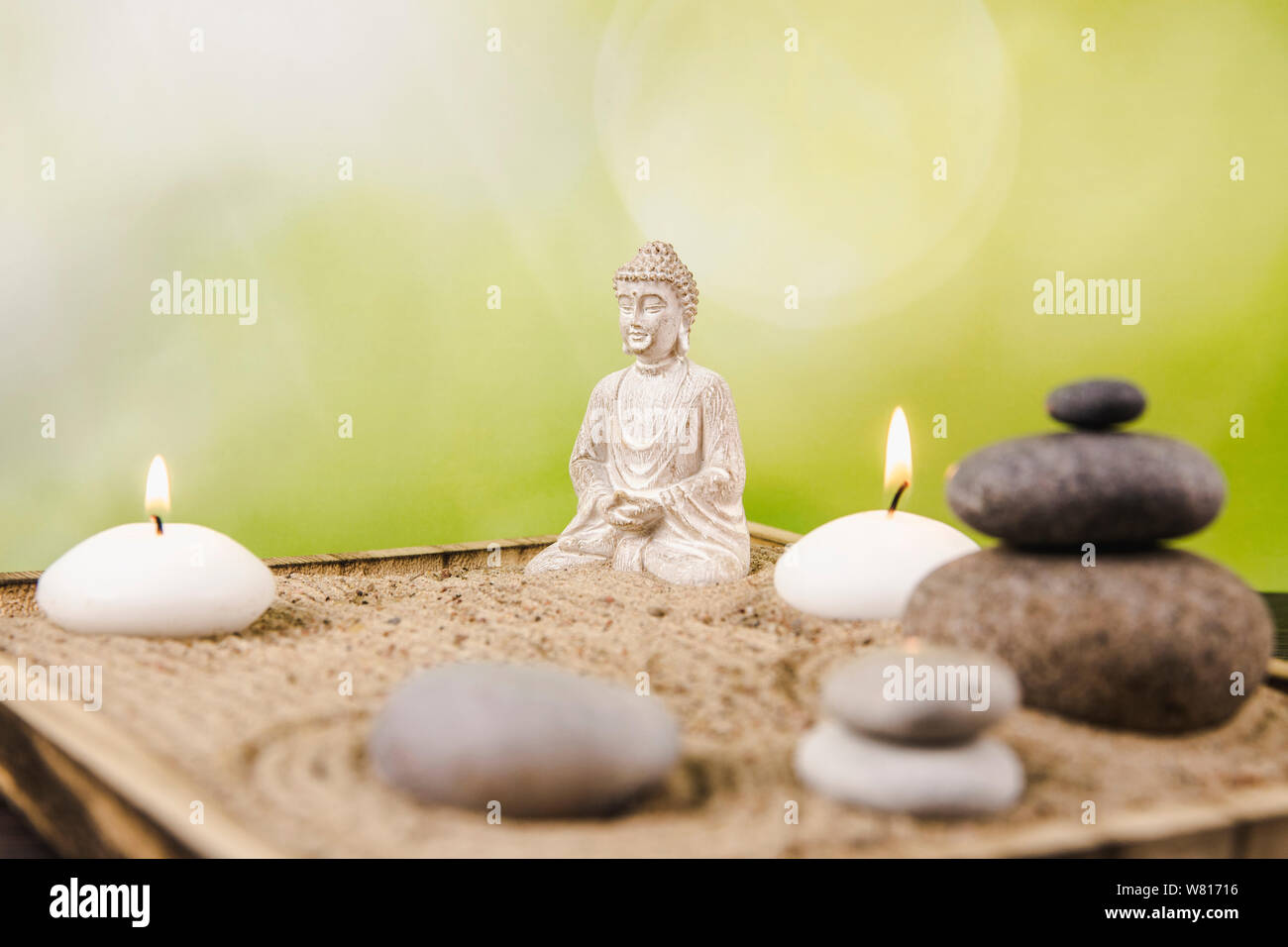 Miniature desk zen sandbox with Buddha figure sit in Lotus position, stacked zen sea stones, spa candles burning against green bokeh studio background Stock Photo
