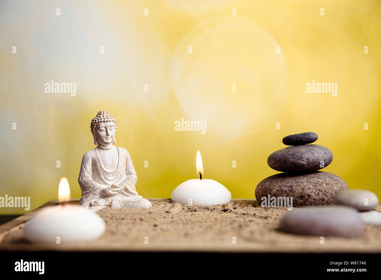 Miniature desk zen sandbox with Buddha figure sit in Lotus position, stacked zen sea stones, spa candles burning against green bokeh studio background Stock Photo