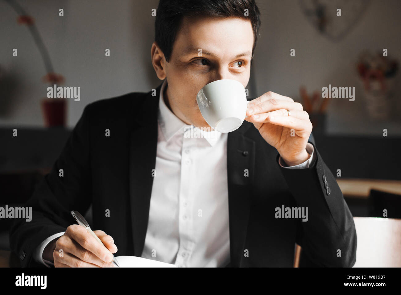 Close up portrait of a confident adult business man writing notes in his daily book while drinking coffee and looking at window . Stock Photo