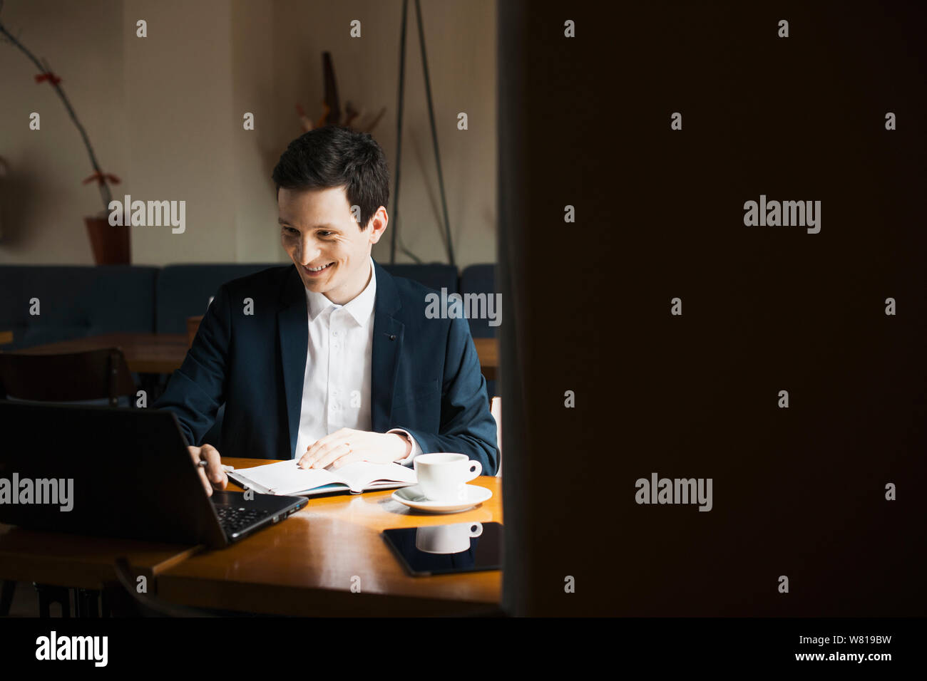 Young stylish manager doing paperwork while operating on his laptop smiling while sitting in a restaurant. Stock Photo
