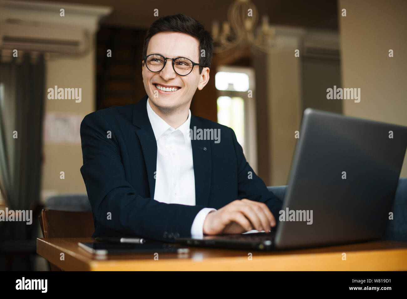 Portrait of a attractive adult freelancer wearing suit and glasses looking away smiling while working at his laptop sitting in a coffee shop. Stock Photo