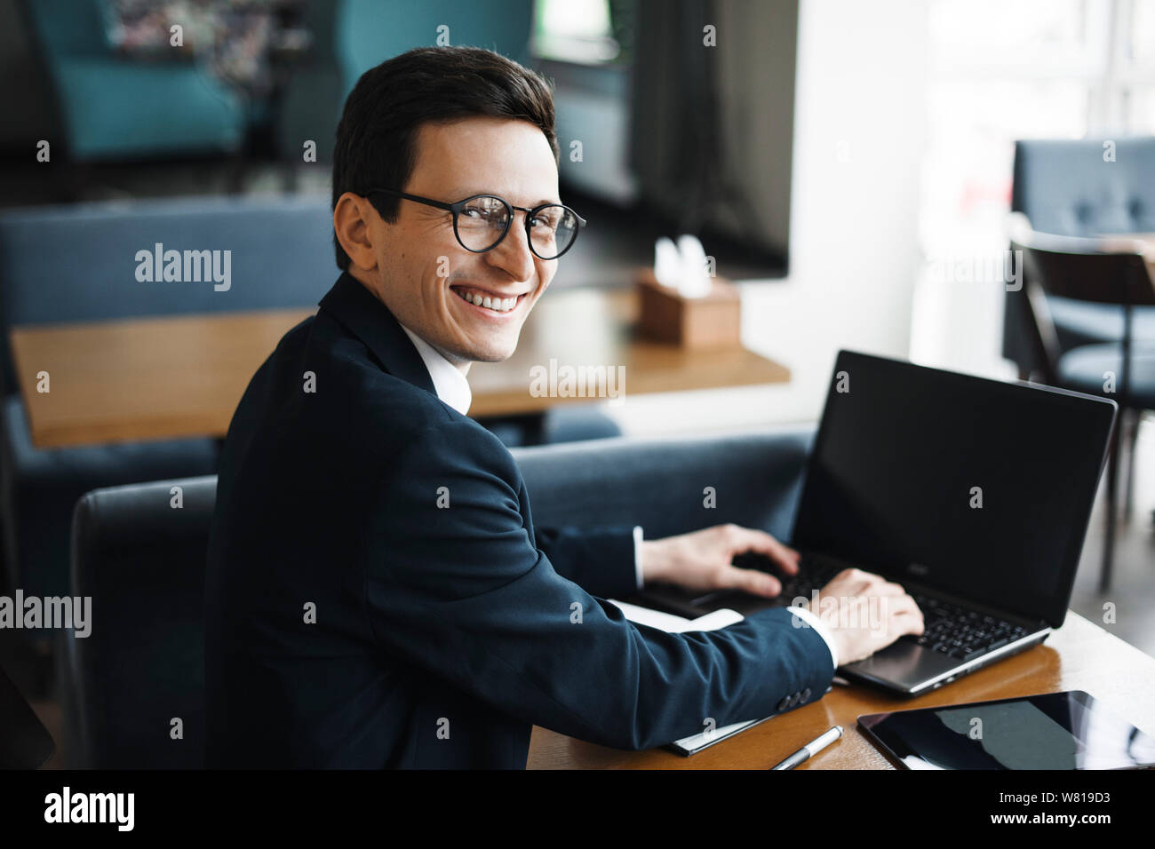 Side view portrait of a stylish adult caucasian manager wearing suit and glasses looking at camera smiling over his shoulder while working at laptop i Stock Photo