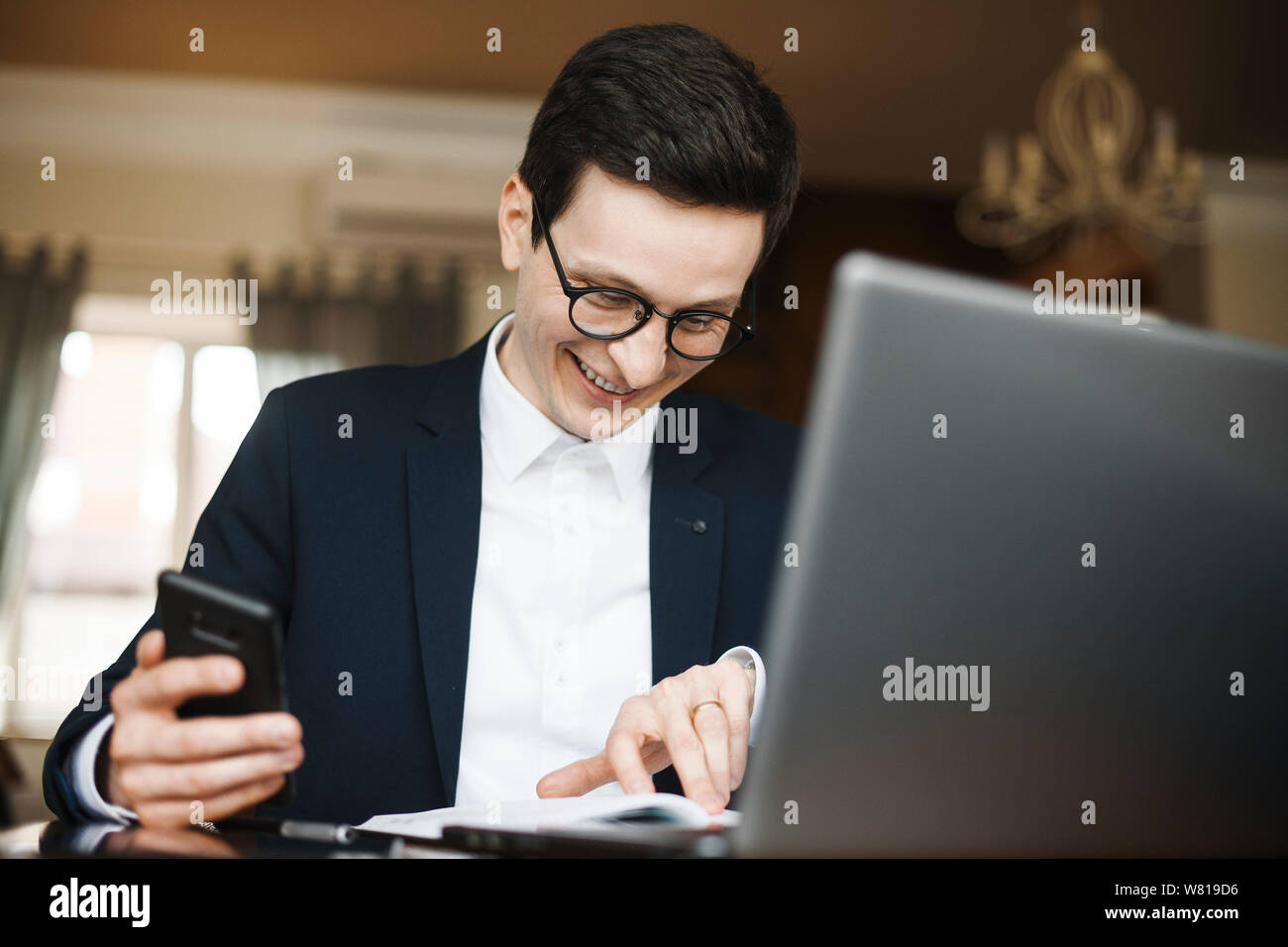 Adult caucasian worker doing paperwork smiling while holding a smartphone in his hand . Stock Photo