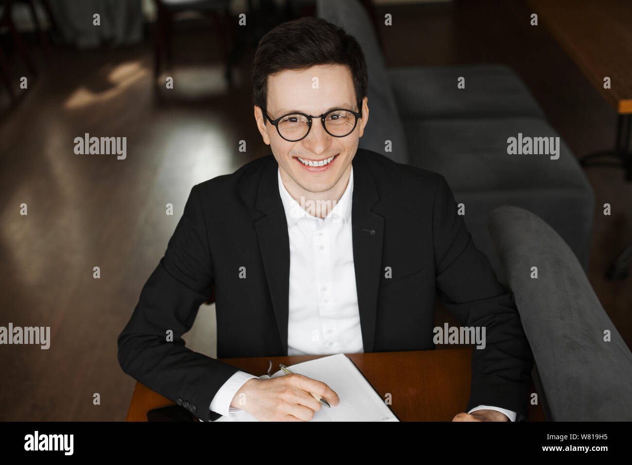 Portrait of a stylish young elegant freelancer looking at camera smiling while sitting at a desk taking notes in his notebook. Stock Photo