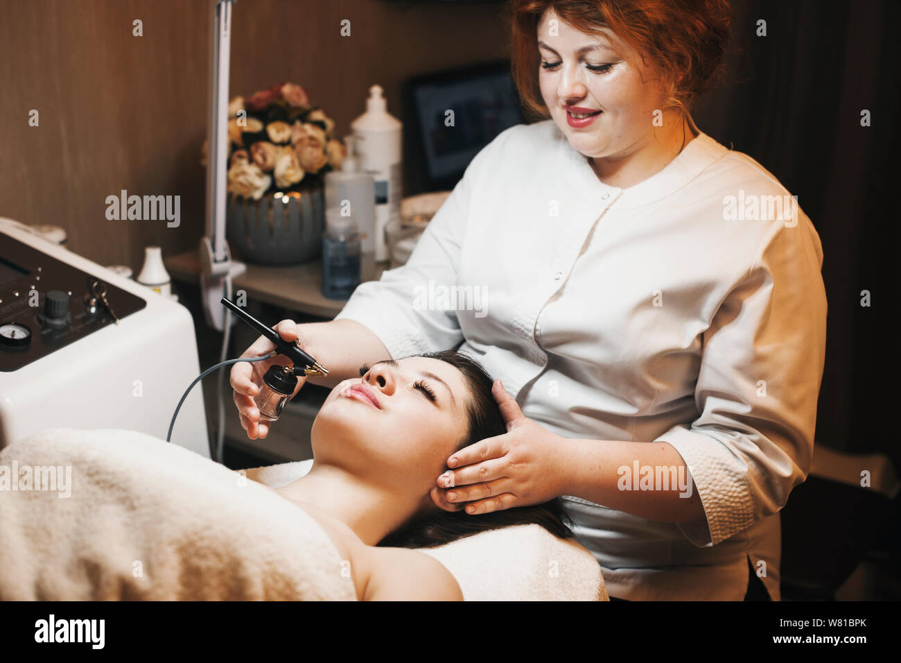 Lovely young female cosmetologist with red hair doing oxygen therapy on a female face in a spa salon. Stock Photo