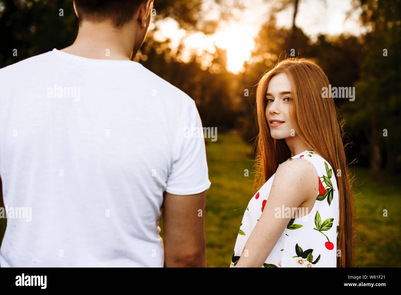 Beautiful red haired woman with freckles looking at camera smiling while holding hand of her boyfriend against sunset while dating outside in the park Stock Photo