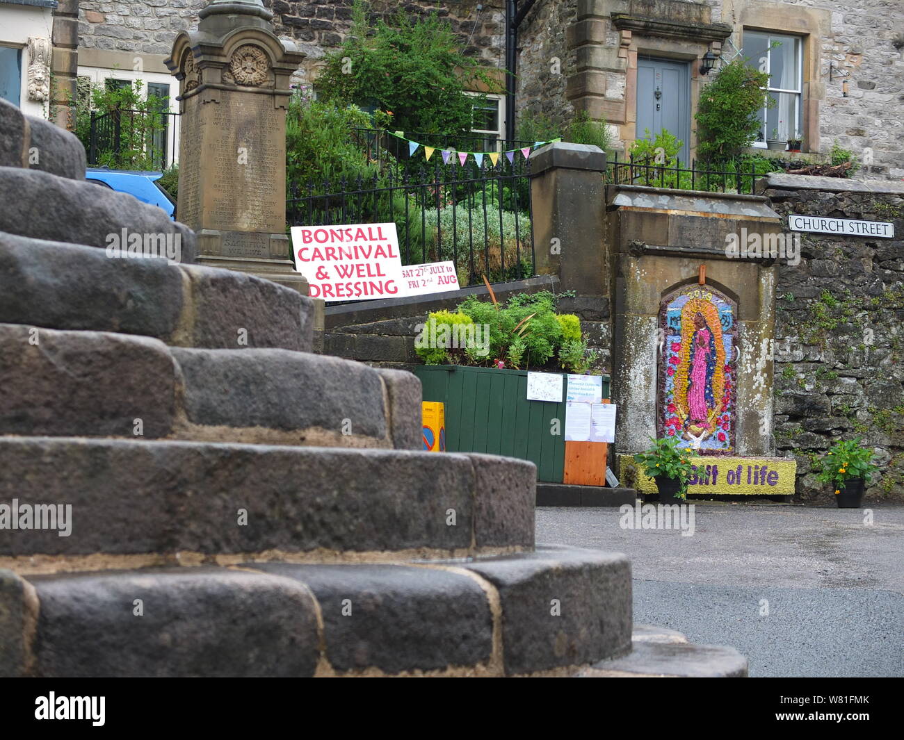 Bonsall a village in the Derbyshire Peak District. The steps of The Cross ancient market cross to foreground with annual well dressing in background. Stock Photo