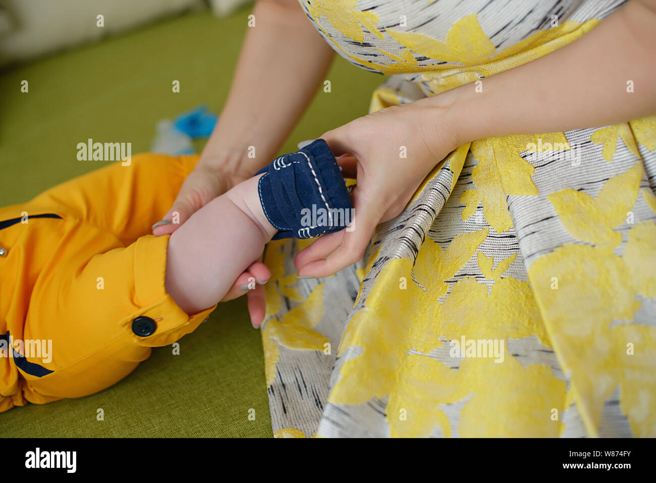 Caucasian young mother wearing a vintage yellow dress and helping little baby boy getting ready, with focus on the hands fitting cute blue booties Stock Photo