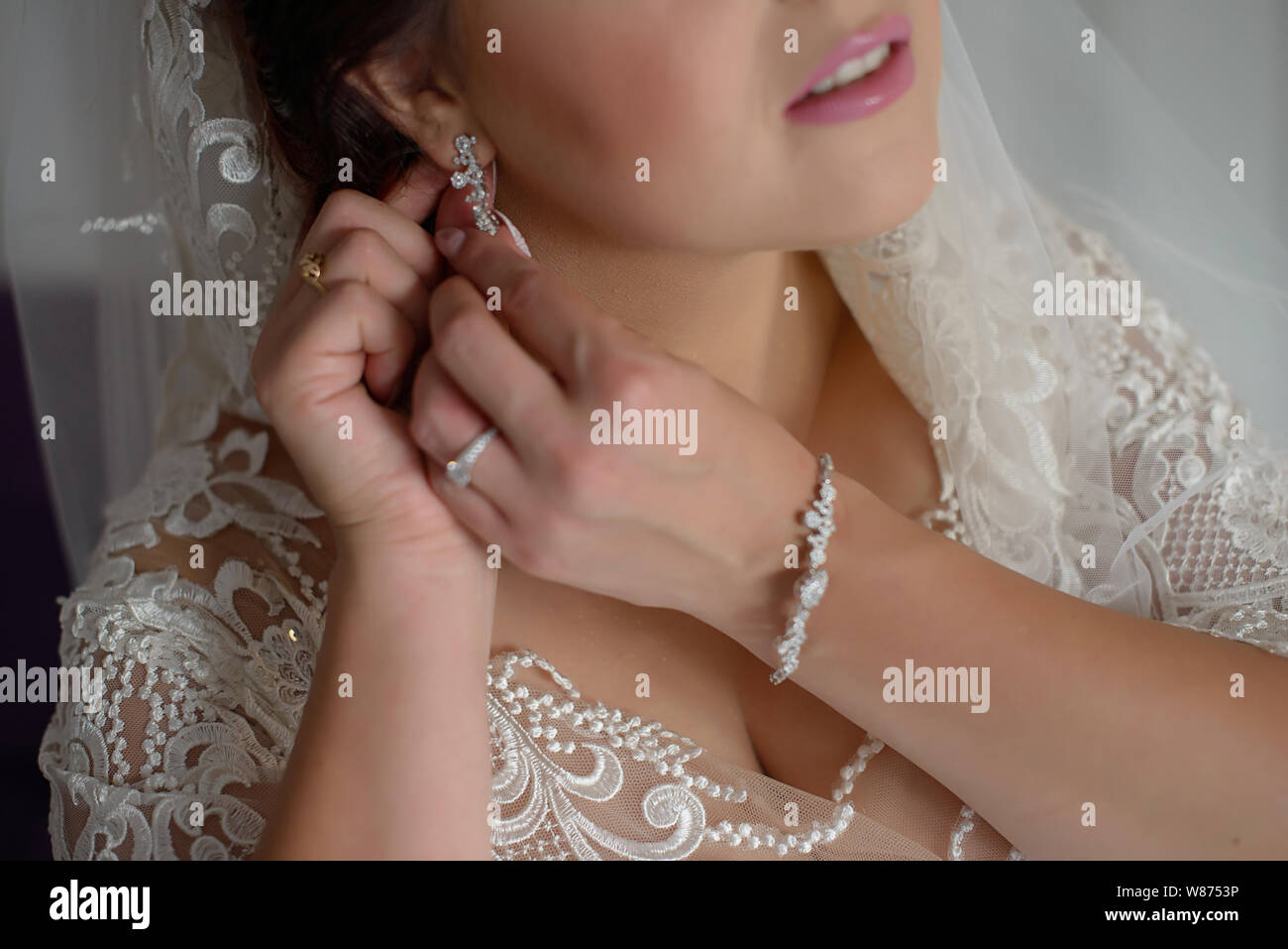 Beautiful bride in white lace wedding dress and embroidered veil wearing vintage diamond jewelry and adjusting earring clasp, getting ready Stock Photo