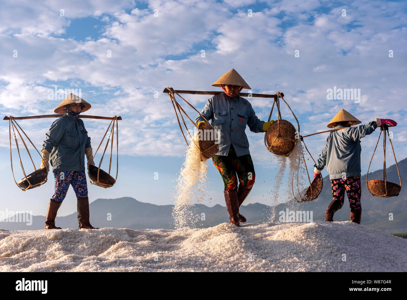 Woman workers pouring freshly harvested salt from baskets to the salt pile at sunrise in Hon Khoi salt field, Nha Trang Province, Vietnam Stock Photo