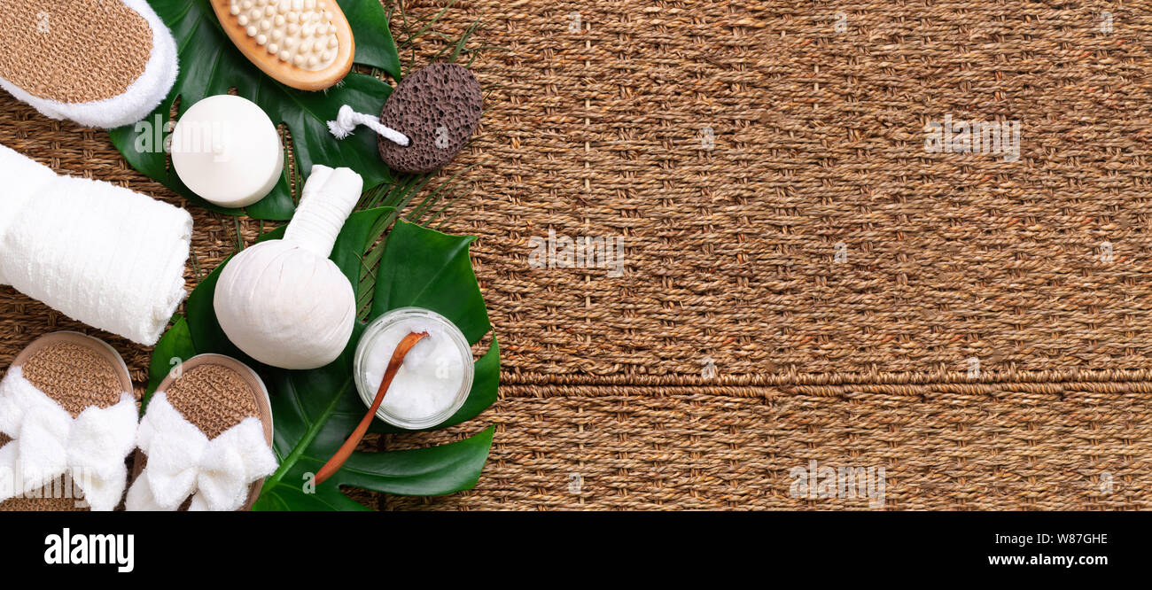 Spa tools: white towel, bamboo slippers, herbal ball, cream, wooden brush, coconut oil, monstera on rattan background. Cosmetic products for body trea Stock Photo