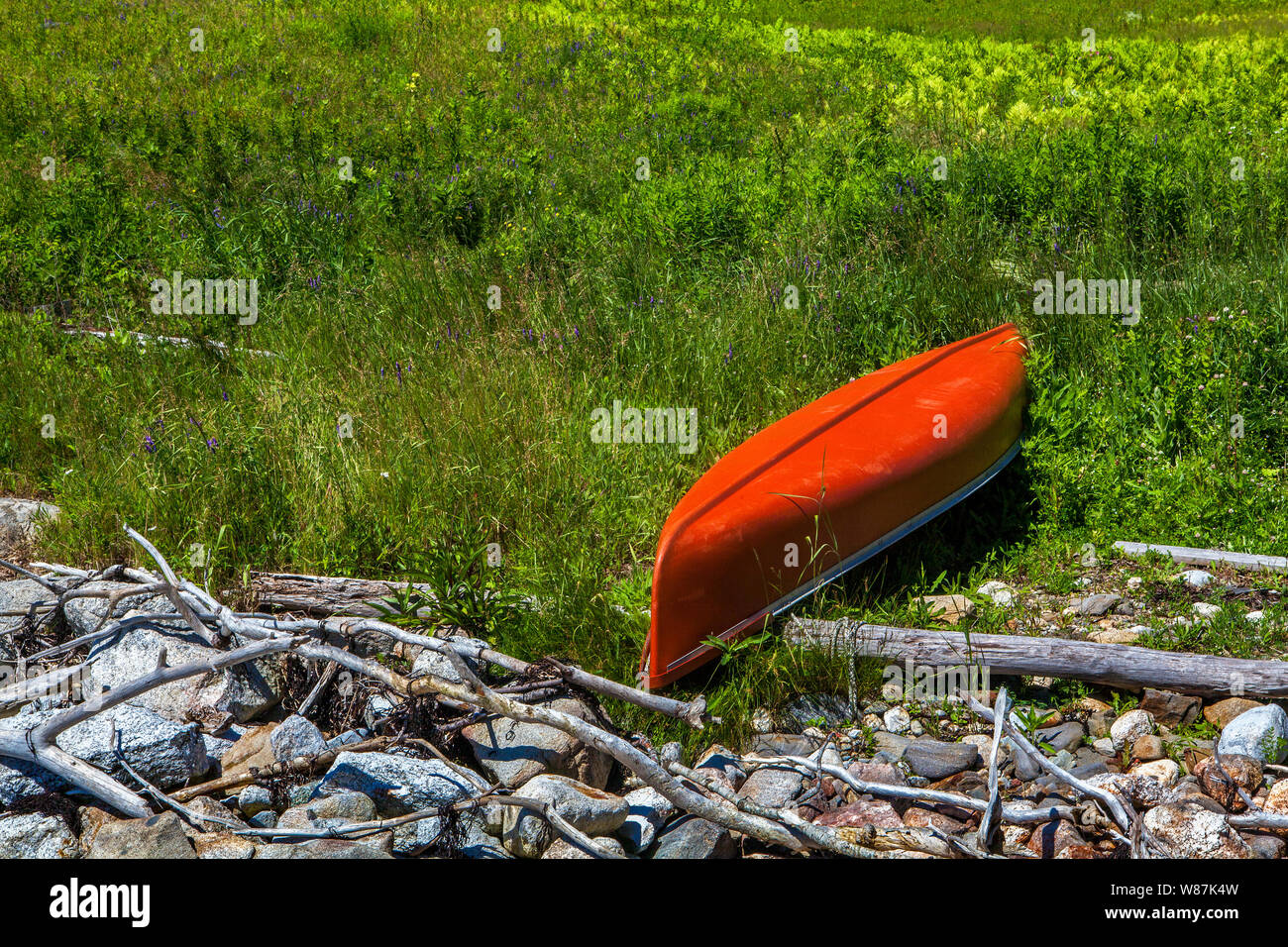 A red-orange upside down canoe sits beside a rocky shorline in a green, grassy field. Stock Photo