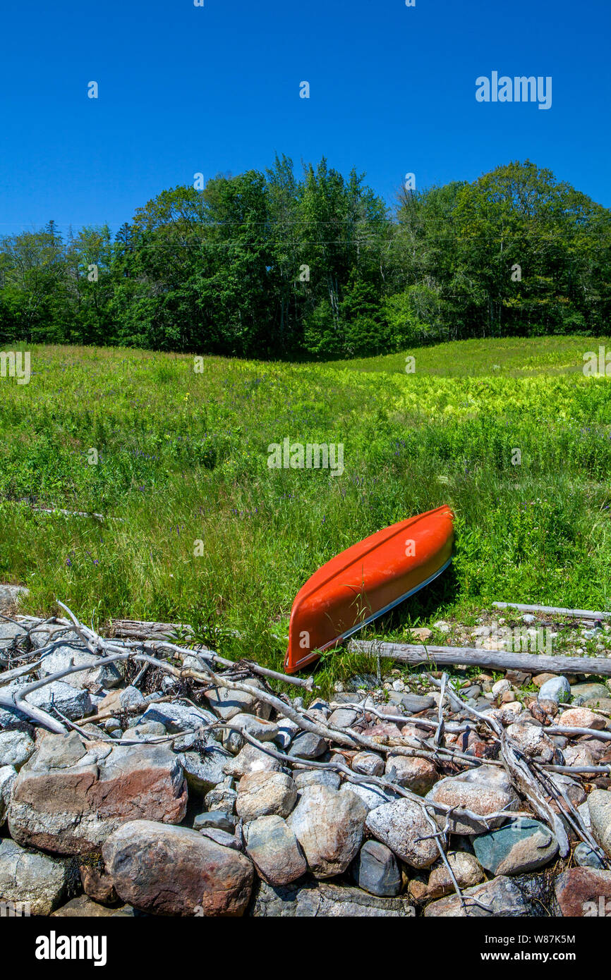 An orange-red upside down canoe beside a rocky shoreline in a green, grassy meadow in Bremen, Maine, USA. Stock Photo